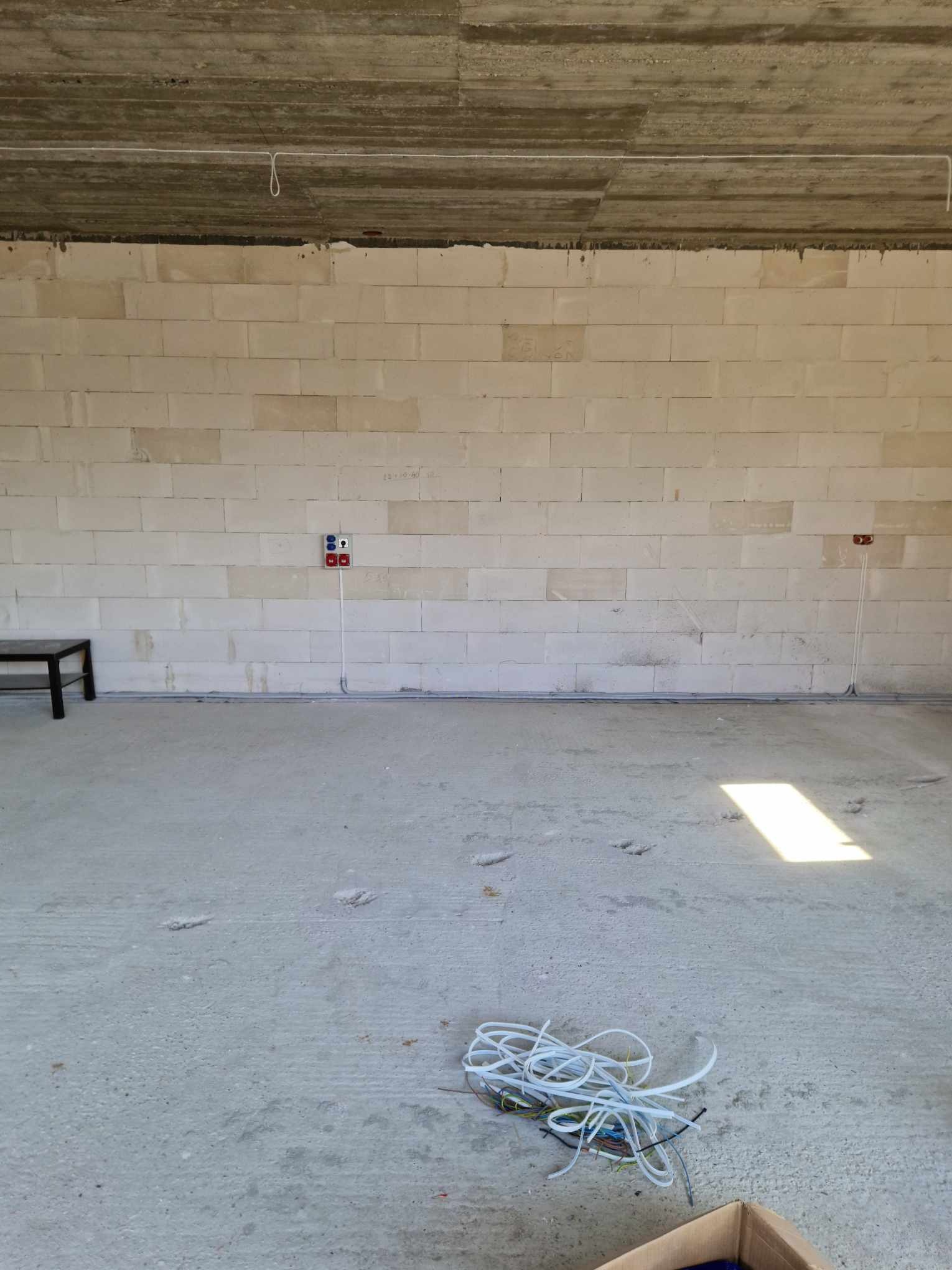 Surowy stan wnętrza z widoczną instalacją elektryczną: kable w peszlach przy ścianie z bloczków, gniazda siłowe, przewody zwisające z surowego stropu, oraz zwój kabli na posadzce.