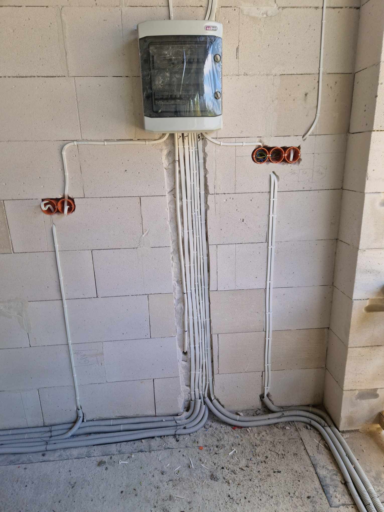 Rozprowadzenie przewodów elektrycznych w ścianie z betonu komórkowego, widoczna skrzynka rozdzielcza w folii ochronnej, peszle przy podłodze oraz puszki instalacyjne w ścianie.