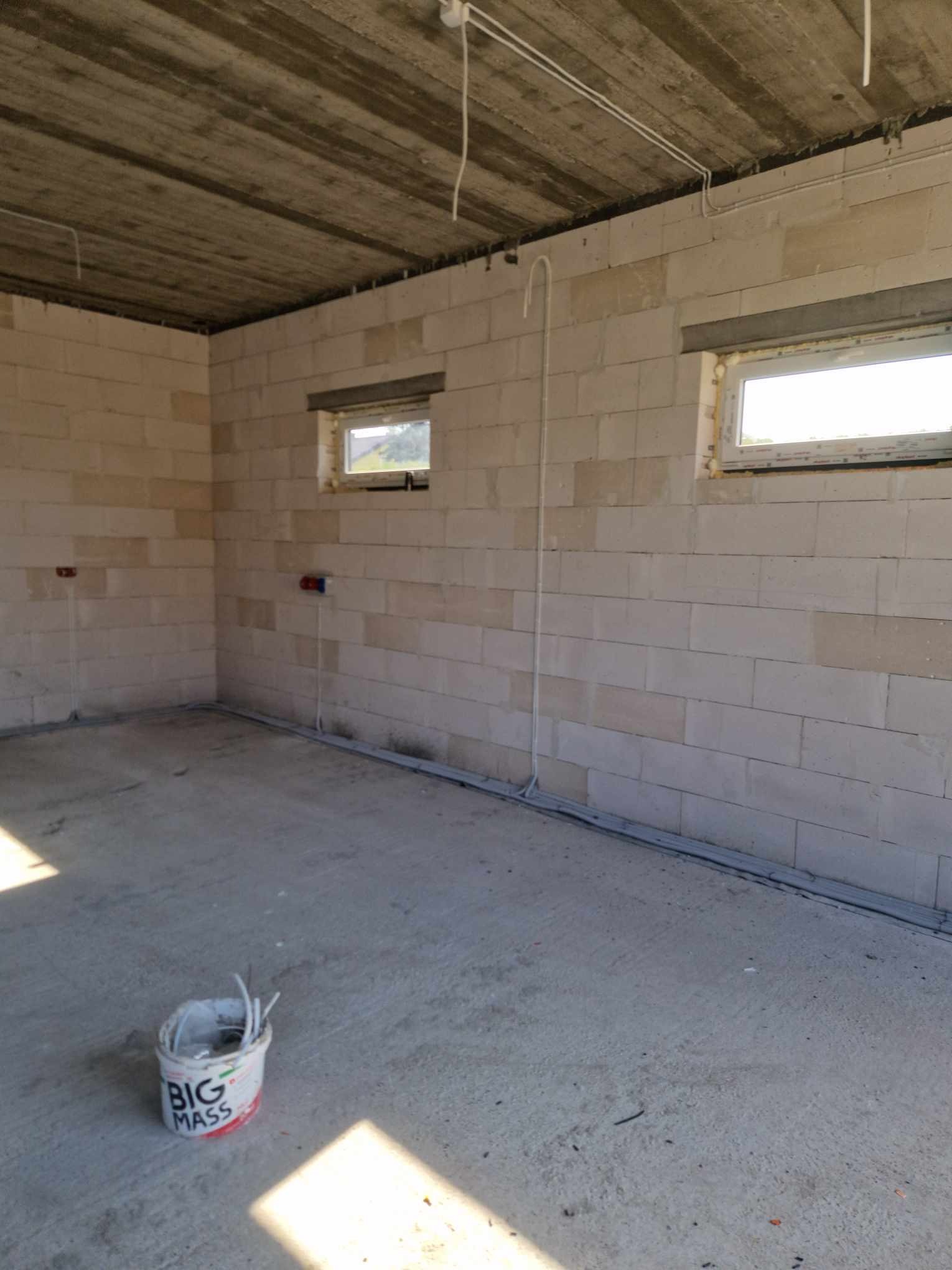 Surowy stan wnętrza z widocznymi przewodami elektrycznymi prowadzonymi po ścianach i suficie, puszki instalacyjne pod oknami, ściany z betonu komórkowego, na podłodze wiadro z narzędziami.