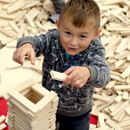 Tomi-Tomi Producent i dystrybutor klocków dla dzieci - Chłopiec w bluzie buduje wieżę z drewnianych klocków Lindenwood, otoczony stertą takich samych klocków.