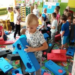 Tomi-Tomi Producent i dystrybutor klocków dla dzieci - Przedszkolaki bawią się klockami piankowymi w kształcie cegieł w sali zabaw z kolorowym dywanem i dekoracjami na ścianach.