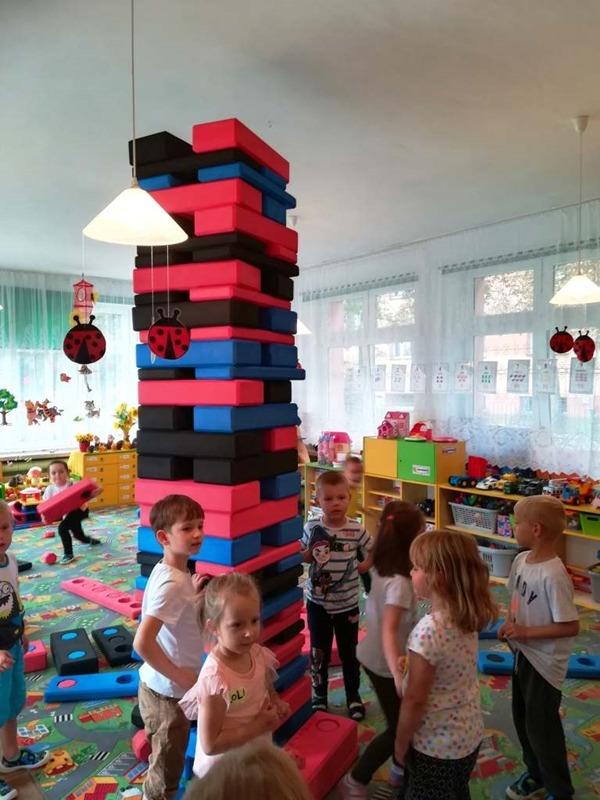 Dzieci w przedszkolu bawią się miękkimi klockami w kolorach czarnym, czerwonym i niebieskim, układając je w wysoką wieżę, podwieszone dekoracje biedronek.