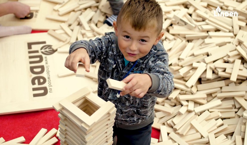 Chłopiec w bluzie buduje wieżę z drewnianych klocków Lindenwood, otoczony stertą takich samych klocków.