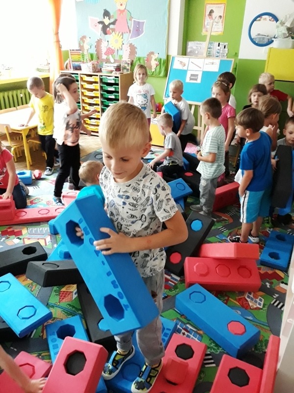 Przedszkolaki bawią się klockami piankowymi w kształcie cegieł w sali zabaw z kolorowym dywanem i dekoracjami na ścianach.