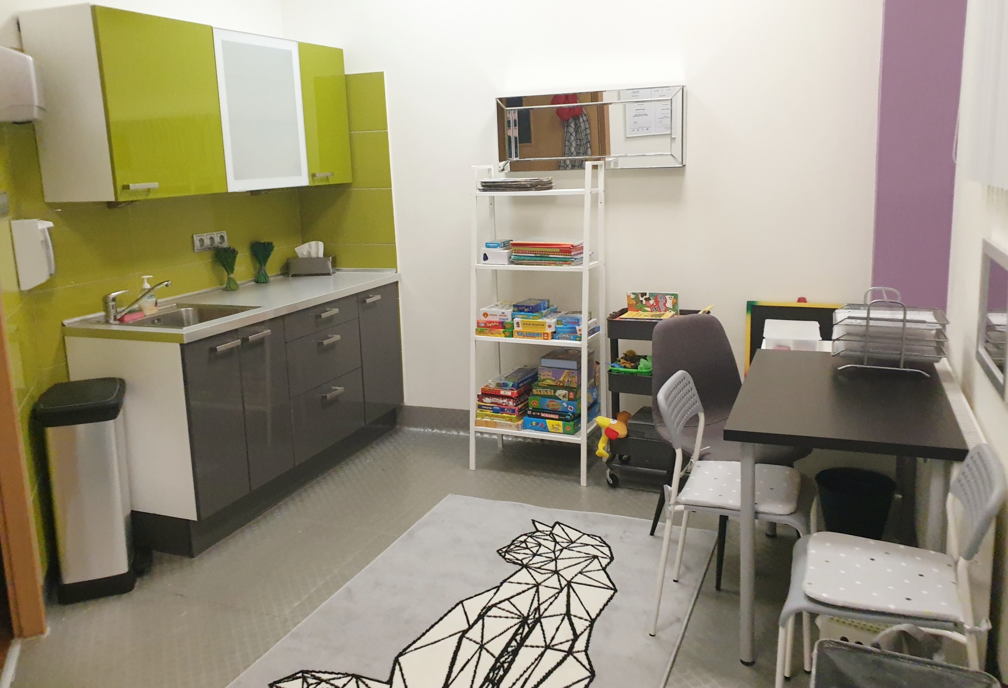 Wnętrze gabinetu z aneksem kuchennym, regałem z grami planszowymi i dywanem z geometrycznym wzorem kota.