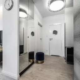 Nowoczesny przedpokój z lustrzaną szafą, okrągłym zegarem ściennym, pufą w kolorze granatowym ze złotą podstawą i okrągłą lampą sufitową z ażurowym kloszem.