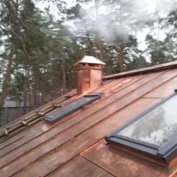 Miedziany dach z oknami dachowymi, kominem i zabezpieczeniami, widok z góry na tle drzew