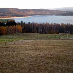 Nowe ogrodzenie z drewnianą bramą na łące z widokiem na jezioro i wzgórza w oddali, w jesiennej scenerii.