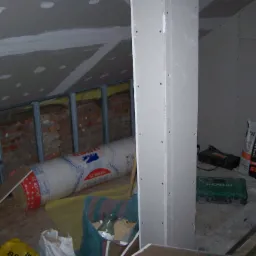 Poddasze w trakcie wykańczania: widoczne płyty gipsowo-kartonowe na ścianach i suficie, izolacja z wełny mineralnej w rolce (Ursa Silver 39) oraz narzędzia budowlane, w tym torba z zaprawą klejową...