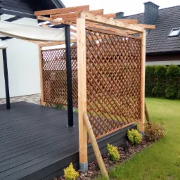Drewniana pergola z ażurową ścianką i rozwijanym zadaszeniem na tarasie z desek kompozytowych, widok na trawnik i dom z szarą dachówką.