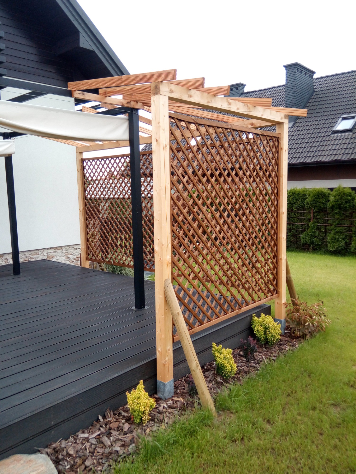 Drewniana pergola z ażurową ścianką i rozwijanym zadaszeniem na tarasie z desek kompozytowych, widok na trawnik i dom z szarą dachówką.