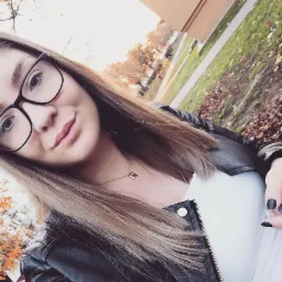 Selfie młodej kobiety w okularach, skórzanej kurtce i z delikatnym naszyjnikiem na tle jesiennego krajobrazu miejskiego.