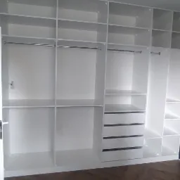 Biała, zabudowana szafa garderobiana z półkami, szufladami i drążkami na ubrania, widoczna na tle parkietu ułożonego w jodełkę i częściowo widocznego łóżka bez materaca.