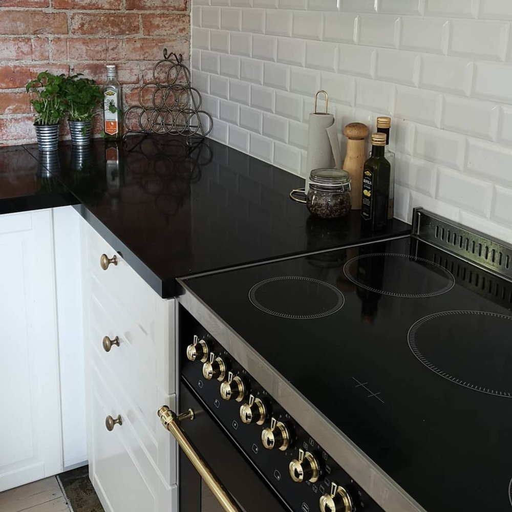 Czarny blat kuchenny z białymi szafkami, złotymi gałkami i płytą indukcyjną, ceglana ściana w tle, detale wystroju.
