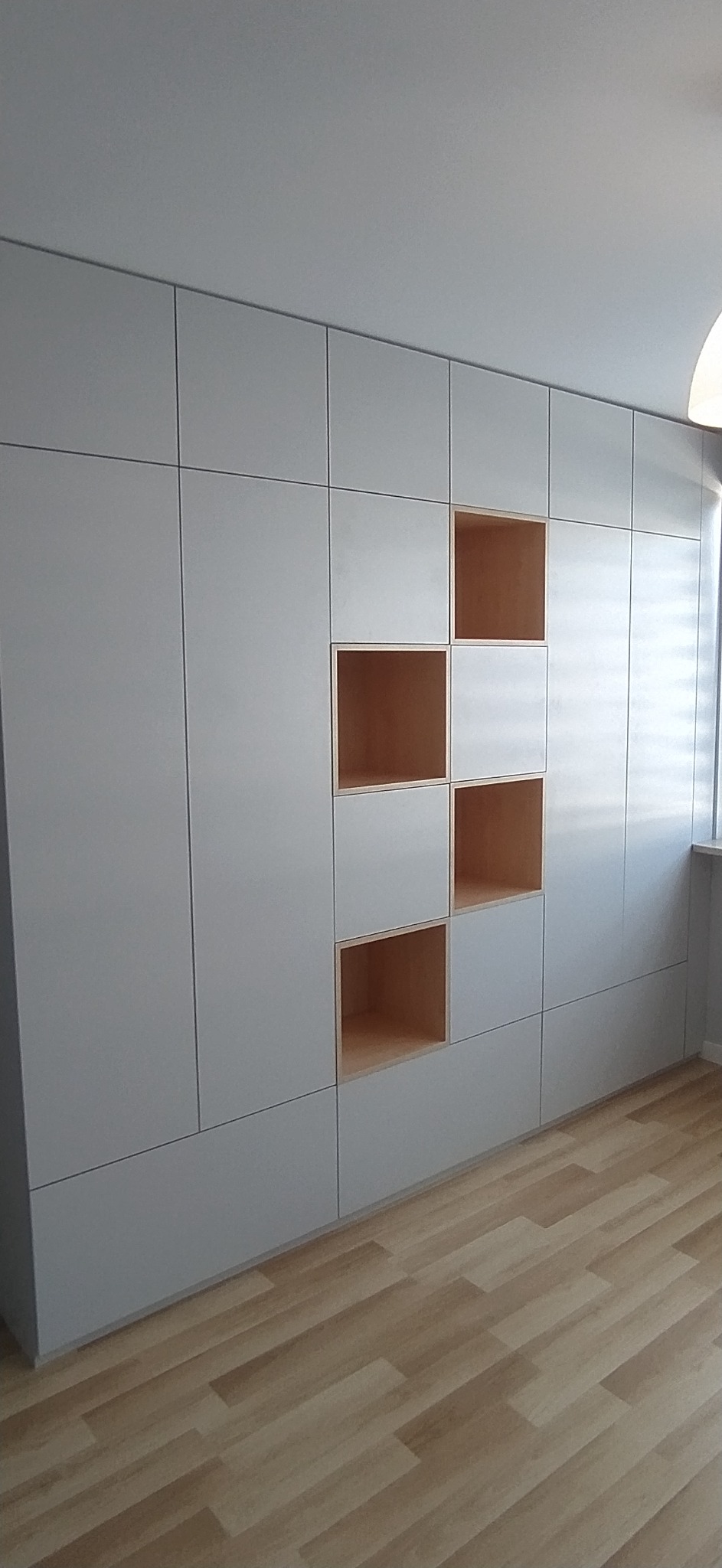 Szara zabudowa meblowa z drewnianymi, otwartymi półkami w nowoczesnym wnętrzu, widoczne panele podłogowe i fragment okna z żaluzjami.
