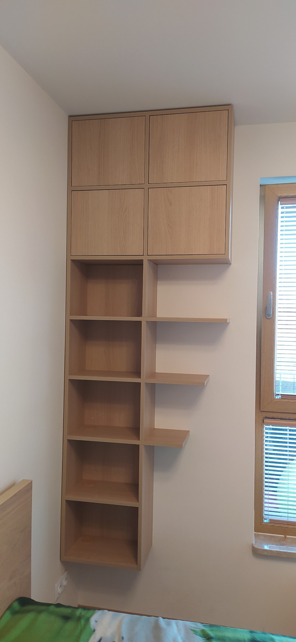 Drewniany regał z szafkami i półkami, zamontowany na ścianie w pokoju, z widocznym fragmentem okna z żaluzjami.