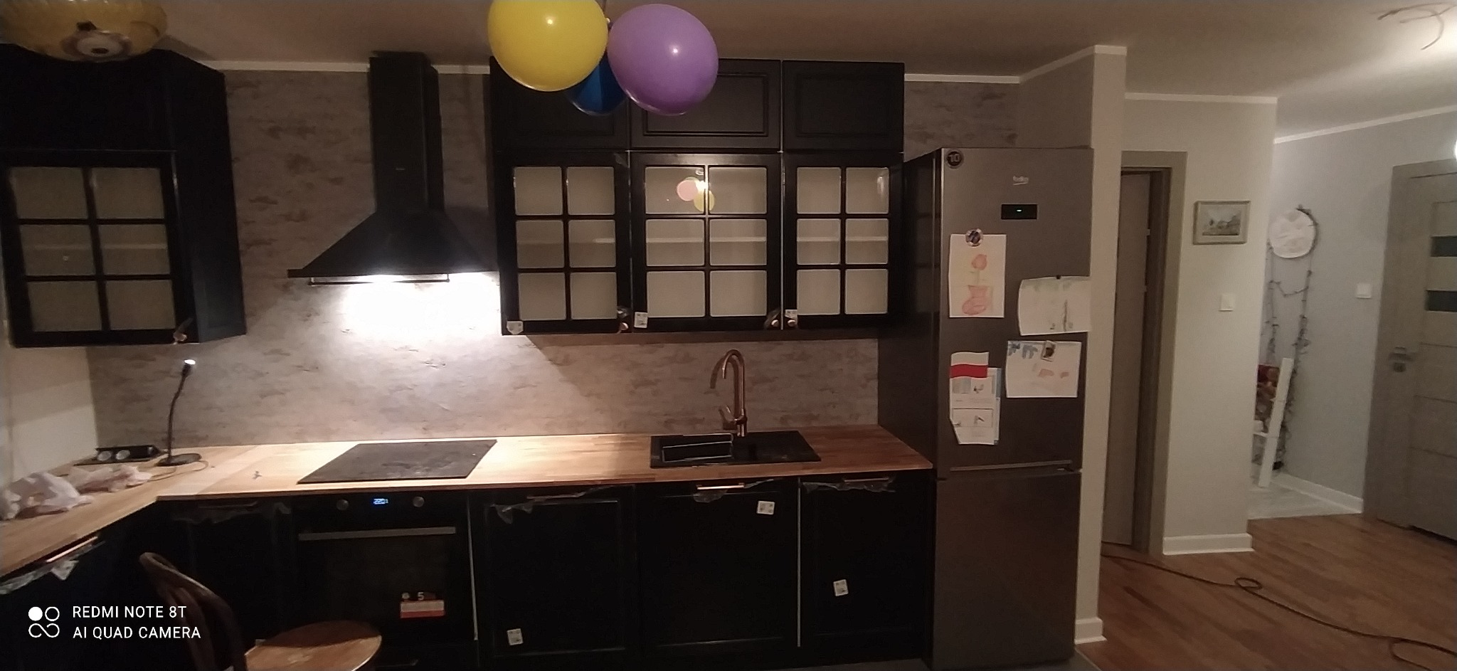 Nowoczesna kuchnia z czarnymi szafkami, drewnianym blatem i miedzianą armaturą, oświetlona punktowo, z balonami dekoracyjnymi wiszącymi pod sufitem.