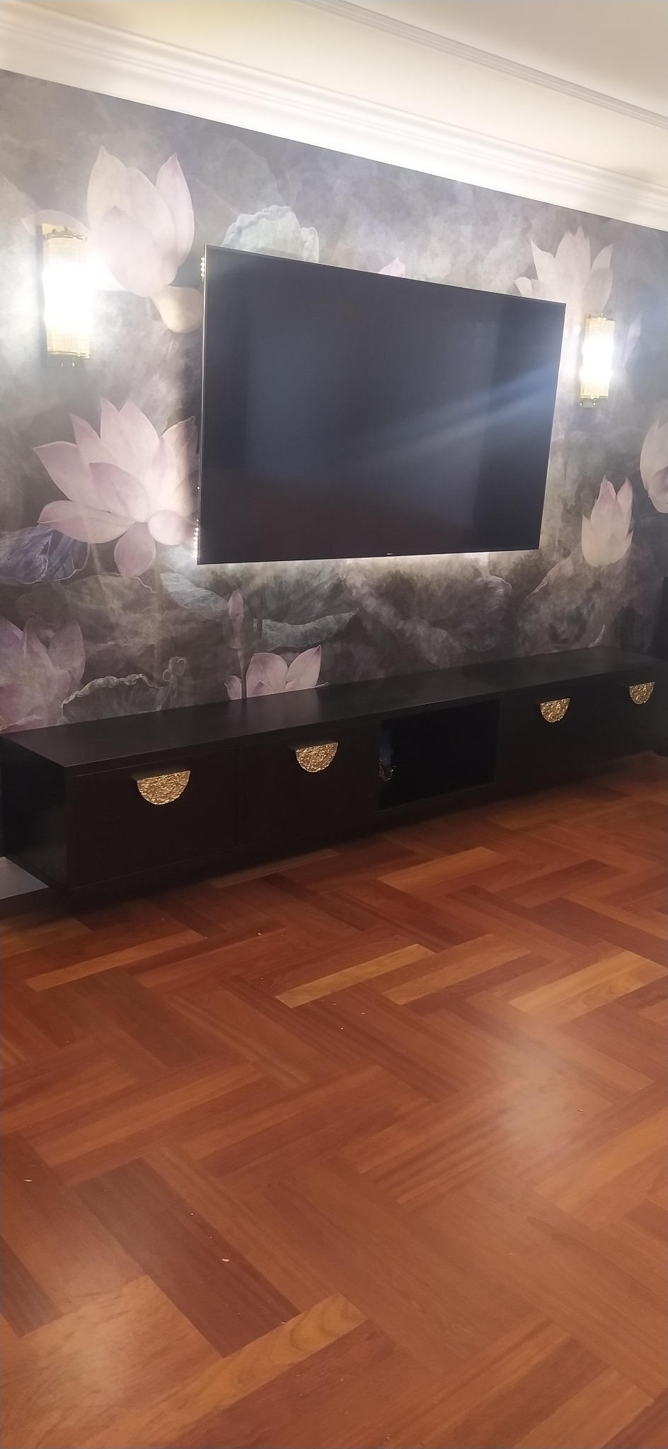Czarna szafka RTV z półokrągłymi, złotymi uchwytami, zamontowana pod telewizorem na ścianie z tapetą w motywy kwiatowe i kinkietami po bokach, parkiet ułożony w jodełkę na podłodze.
