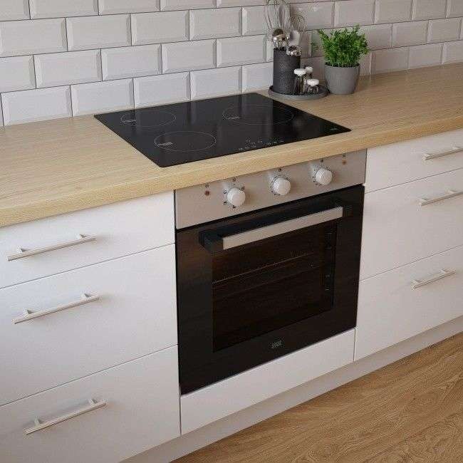 Nowoczesna kuchnia z białymi szafkami, drewnianym blatem, płytą indukcyjną i piekarnikiem z srebrnymi pokrętłami, uzupełniona białymi płytkami na ścianie i ziołami w doniczce.