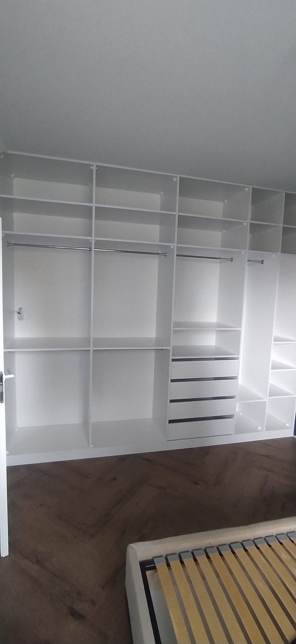 Biała, zabudowana szafa garderobiana z półkami, szufladami i drążkami na ubrania, widoczna na tle parkietu ułożonego w jodełkę i częściowo widocznego łóżka bez materaca.