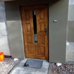 Drewniane drzwi wejściowe po remoncie, osadzone w świeżo otynkowanym portalu, widoczny fragment chodnika z szarej kostki brukowej i pomarańczowe wiadro w rogu.