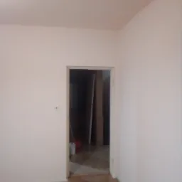 Pomalowane na jasny róż ściany pokoju z drewnianą podłogą, widok na otwarte drzwi do innego pomieszczenia, gdzie widać wiadro i materiały budowlane.