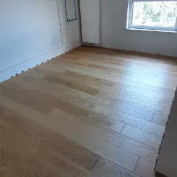 Nowo położona, jasna drewniana podłoga w pokoju z białymi ścianami, widoczne podkładki dystansowe przy ścianach, okno z roletą i grzejnik.