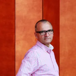 Portret mężczyzny w okularach i różowej koszuli na tle panelowej ściany w odcieniach pomarańczy, prawdopodobnie doradcy finansowego z Koszalina.