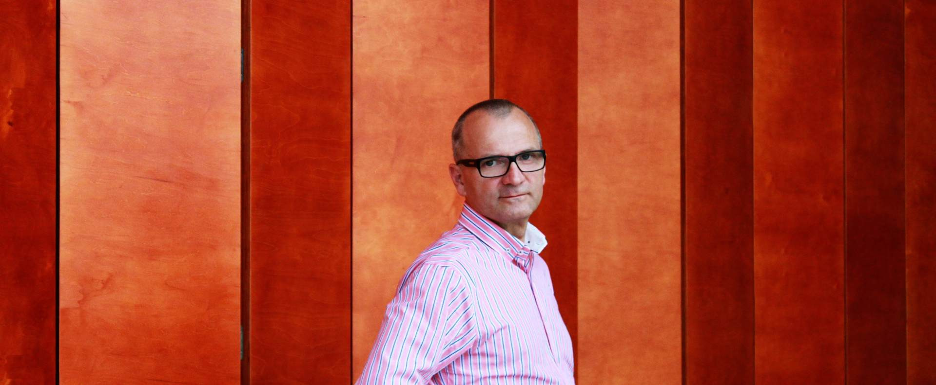 Portret mężczyzny w okularach i różowej koszuli na tle panelowej ściany w odcieniach pomarańczy, prawdopodobnie doradcy finansowego z Koszalina.