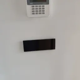 Panel alarmowy Satel Perfecta i czarny szklany panel dotykowy z trzema niebieskimi diodami LED zamontowane na gładkiej, białej ścianie.