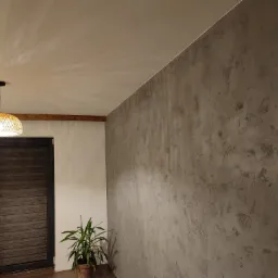 Ściana wykończona dekoracyjnym tynkiem w odcieniach szarości, oświetlona wiszącą lampą z abażurem z naturalnego materiału, widoczna drewniana belka pod sufitem, drzwi z poziomymi panelami...