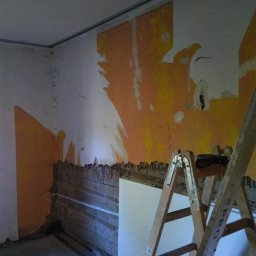 VOLT Bartłomiej Motyka - Pokój w trakcie przygotowania do prac wykończeniowych: zdarte fragmenty farby na ścianach, odsłonięte deski, drabina oparta o ścianę, worki z zaprawą na podłodze.