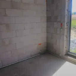 Surowy pokój z bloków betonowych z widocznymi przewodami elektrycznymi, puszkami instalacyjnymi i wylewką podłogową, okno z widokiem na zieleń na zewnątrz.