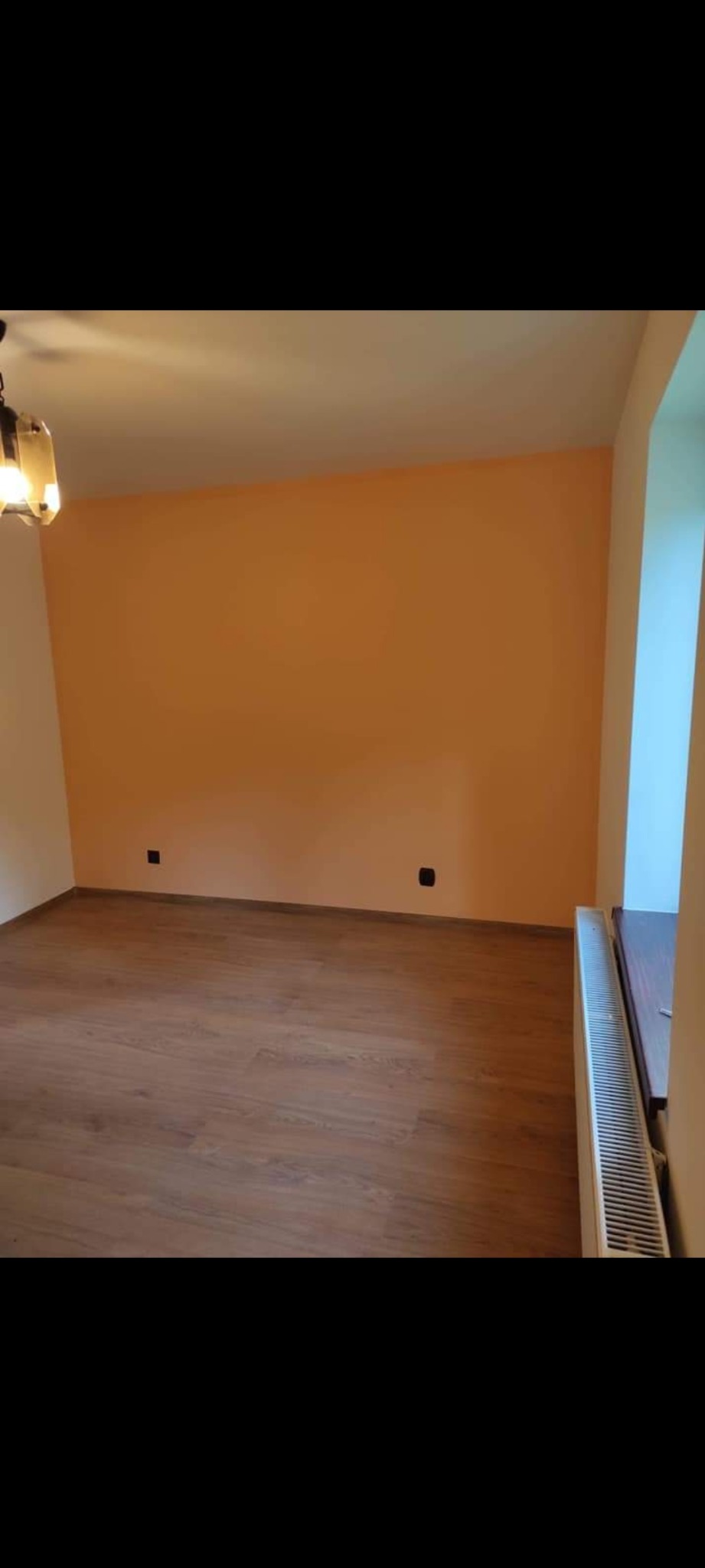 Pomalowane na pomarańczowo wnętrze pokoju z drewnianą podłogą, białym grzejnikiem i lampą sufitową w rogu.