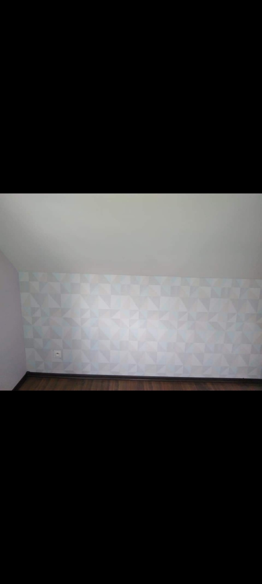 Ściana z geometryczną tapetą w odcieniach szarości i błękitu, widoczny fragment podłogi z drewnianymi panelami i biały sufit. Na ścianie gniazdko elektryczne.