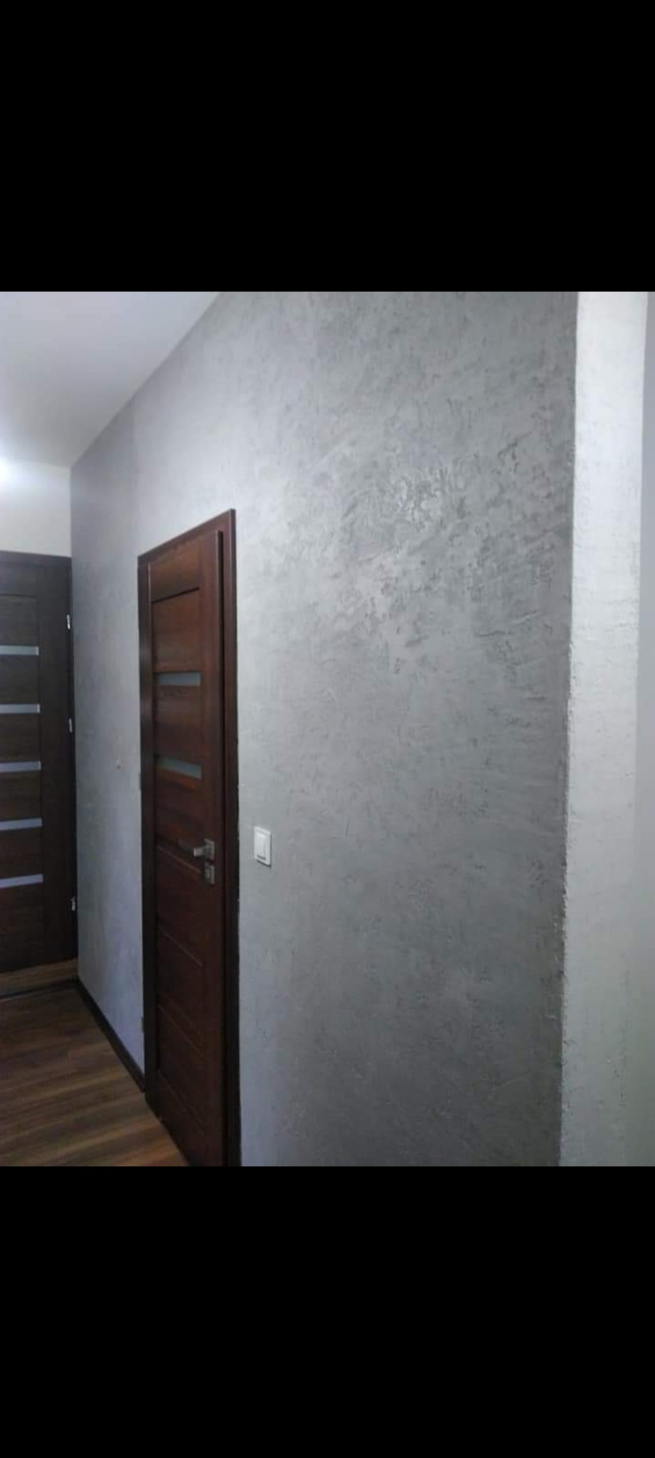 Ściana w korytarzu pokryta dekoracyjną szarą farbą z efektem betonu, widoczne dwie drewniane drzwi z przeszkleniami i włącznik światła.