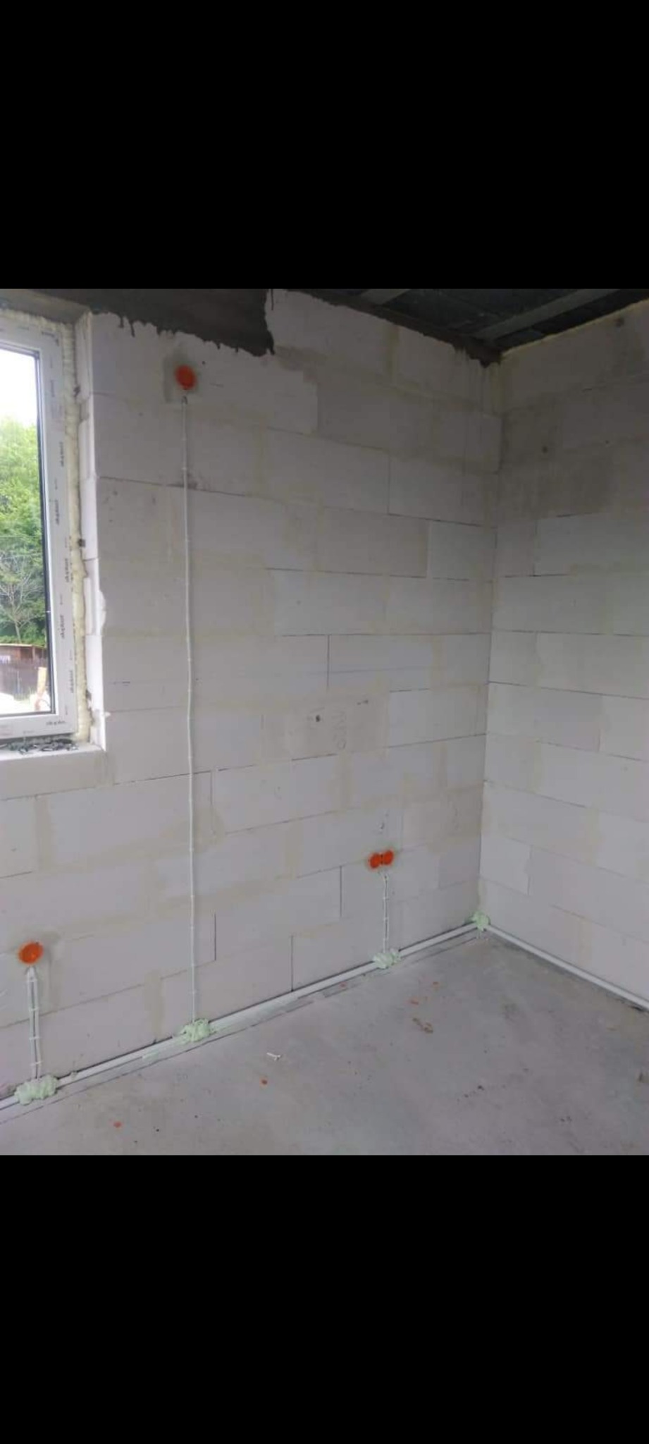 Surowy stan wnętrza z białych bloczków, widoczne pomarańczowe puszki elektryczne i białe przewody przytwierdzone pianką montażową, okno z widokiem na zieleń.