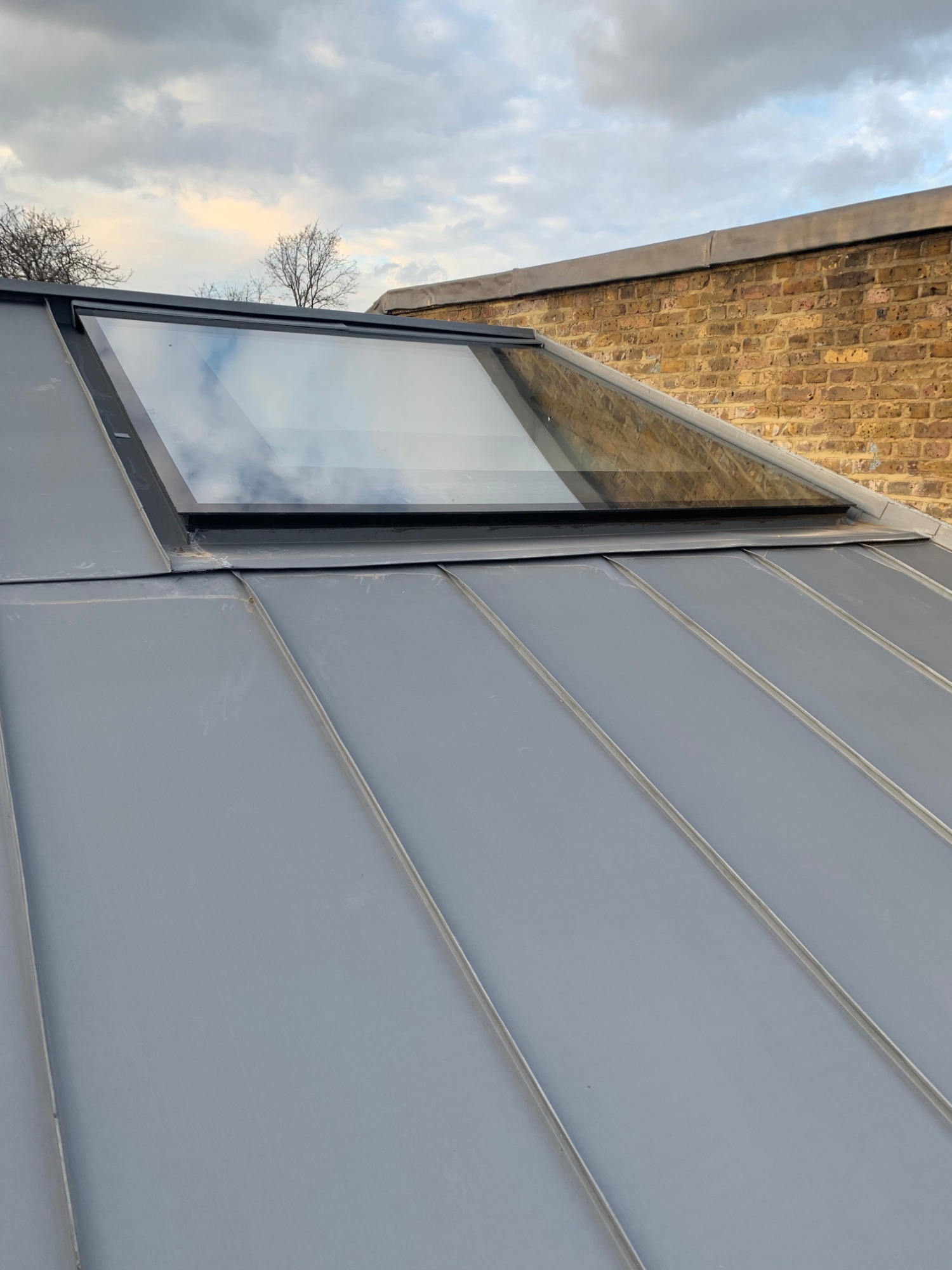 Szary dach z blachy na rąbek stojący z oknem dachowym, odbijającym pochmurne niebo, na tle ceglanego budynku.