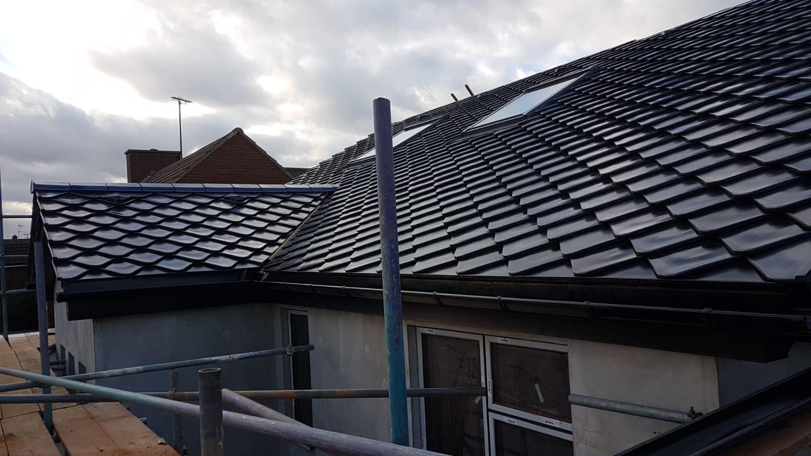 Nowy, błyszczący dach z czarnej dachówki karpiówki z oknem dachowym, widoczny z poziomu rusztowania. W tle fragment dachu sąsiedniego budynku.