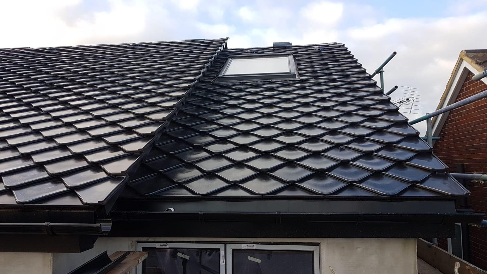 Błyszczący, czarny dach z dachówki karpiówki z oknem dachowym i zamontowanym orynnowaniem, widoczne rusztowanie i antena telewizyjna w tle.