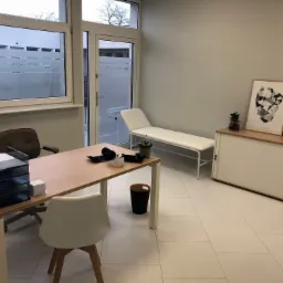 Jasne wnętrze gabinetu lekarskiego z biurkiem, kozetką i dekoracjami, widoczne logo Medycyna Pracy na szybie okna.