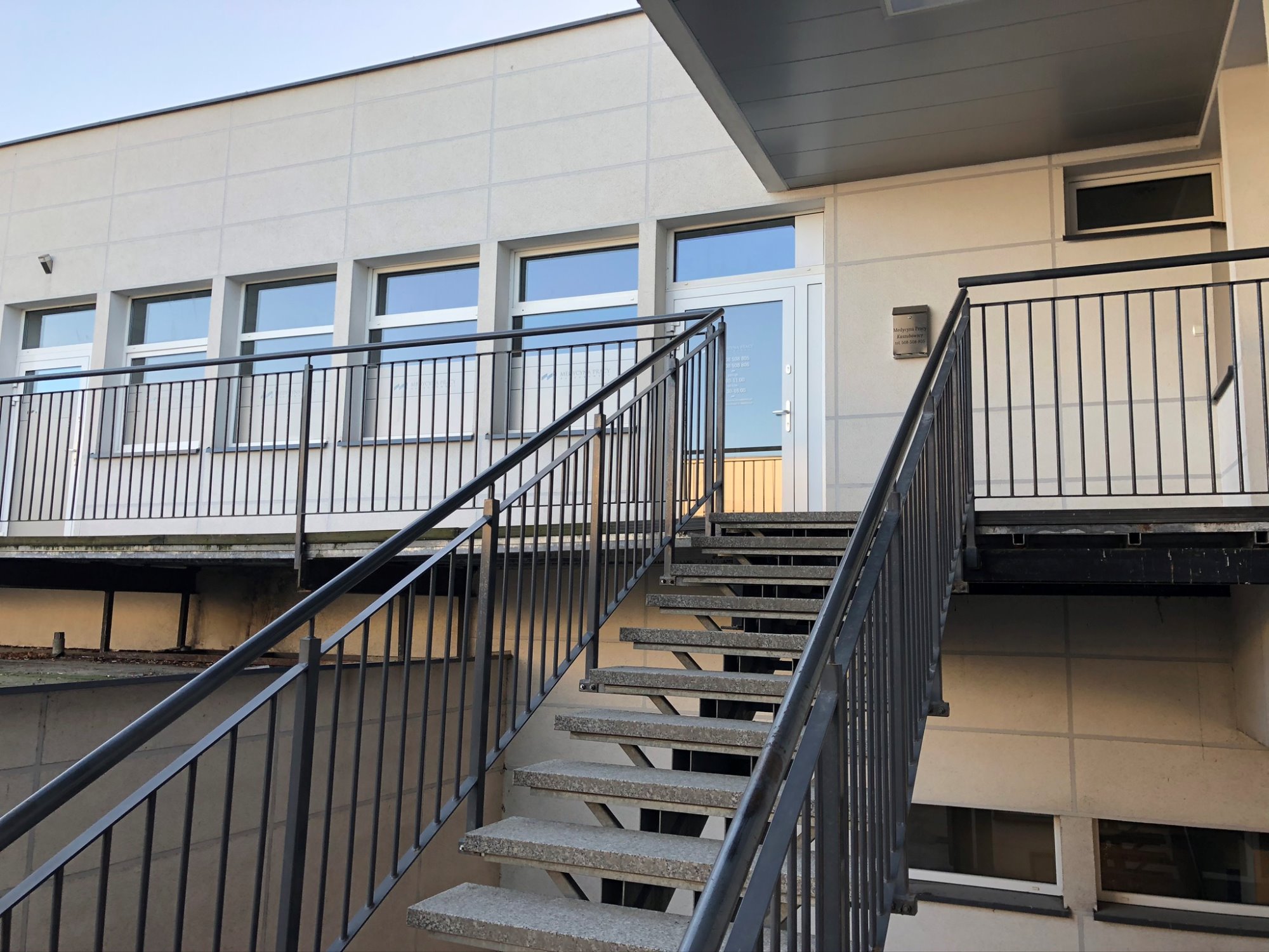Wejście do budynku z metalowymi schodami i balustradami, tabliczka informacyjna Medycyny Pracy przy drzwiach.