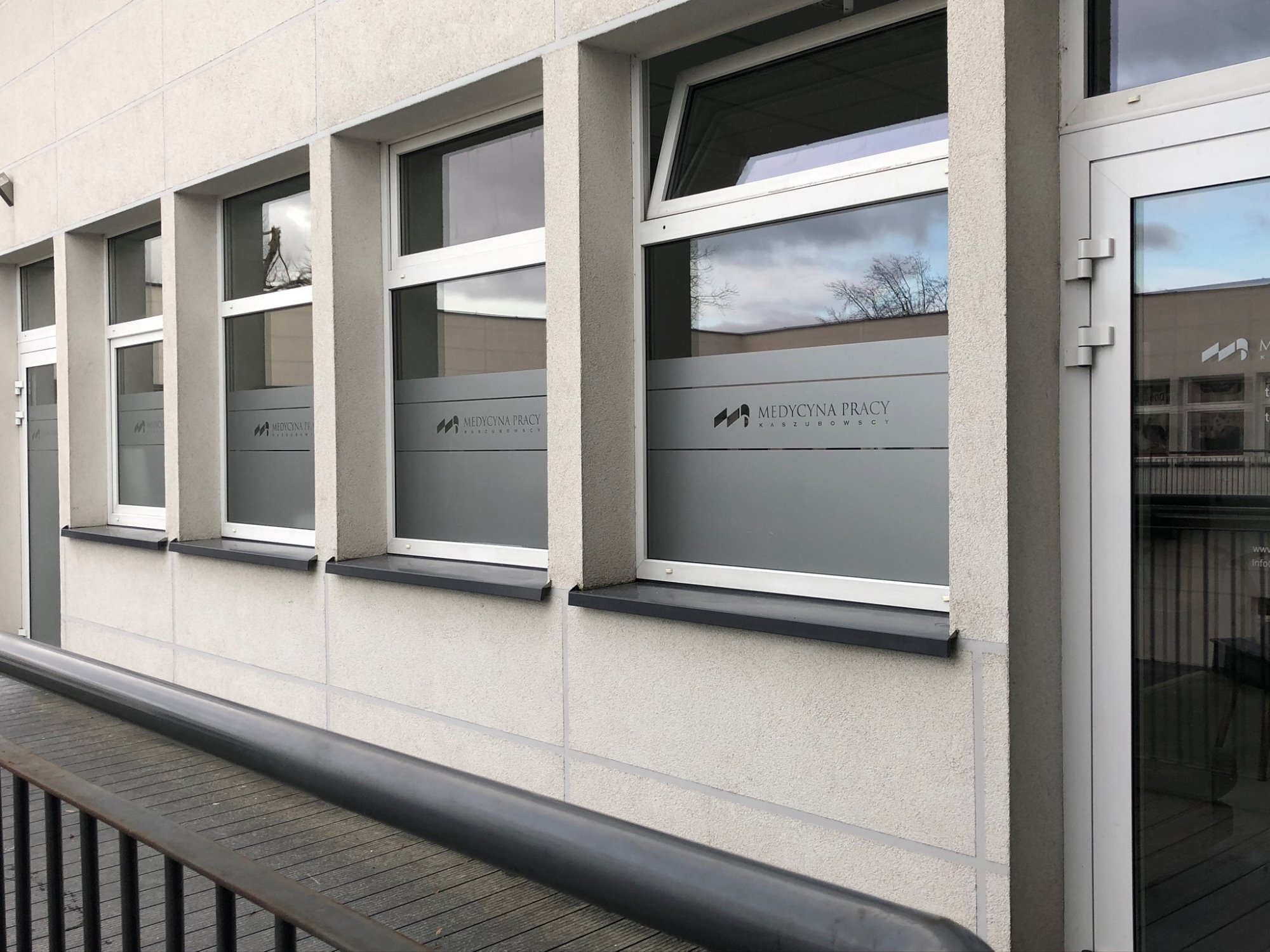 Fasada budynku z oknami z naklejonym logo i napisem 'Medycyna Pracy Kaszubscy', widoczny podjazd dla niepełnosprawnych.