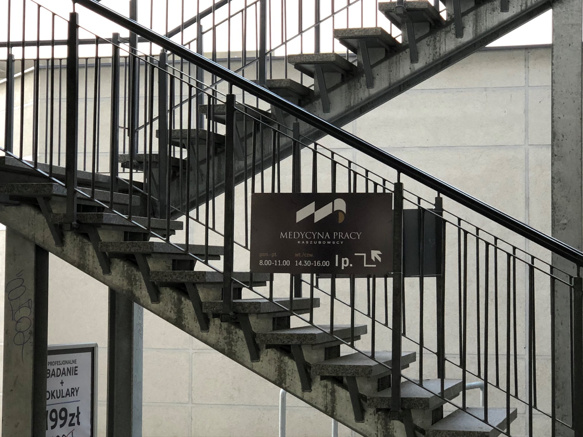 Tablica informacyjna 'Medycyna Pracy Kaszubowscy' umieszczona na tle schodów z metalową balustradą i betonowymi stopniami, widok z dołu.