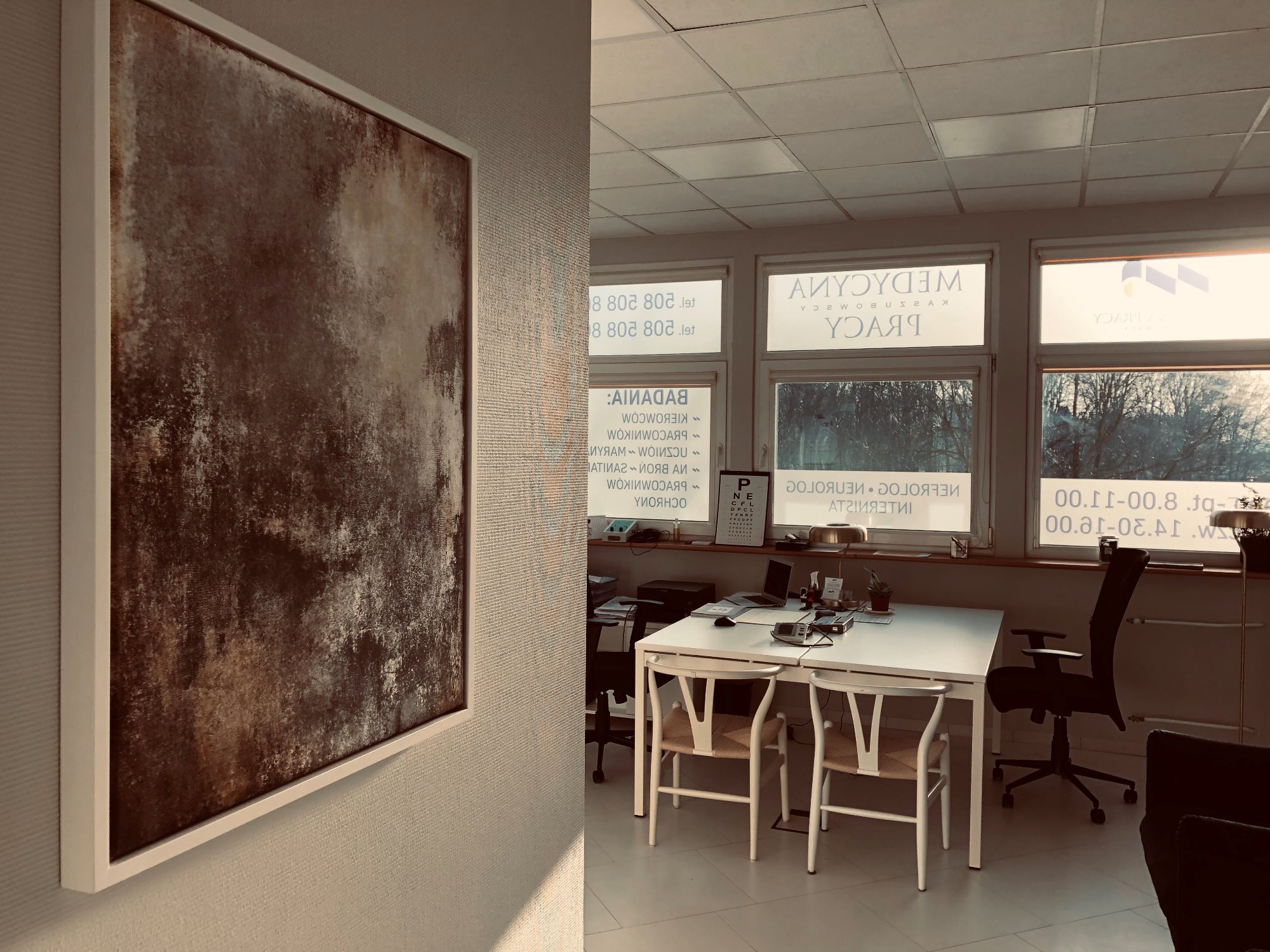 Wnętrze gabinetu Medycyny Pracy Chojnice z widocznym obrazem na ścianie, biurkami i krzesłami, oraz naklejkami na oknach z informacjami o firmie i godzinach pracy.