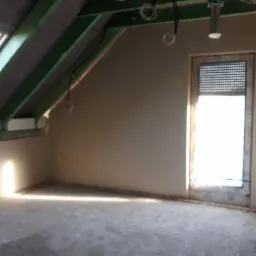 Poddasze w trakcie prac wykończeniowych: ściany i podłoga pokryte świeżym tynkiem, widoczne drewniane elementy konstrukcji dachu i okno dachowe.