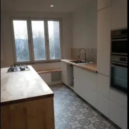 Jasna kuchnia z drewnianym blatem, białymi szafkami, wzorzystą podłogą i wnęką z siedziskiem pod oknem; widoczne niedokończone podłączenie oświetlenia sufitowego.