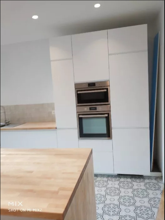 Nowoczesna kuchnia z białymi szafkami, zabudowanym piekarnikiem i płytą, drewnianym blatem wyspy kuchennej oraz podłogą w szaro-białe wzory.