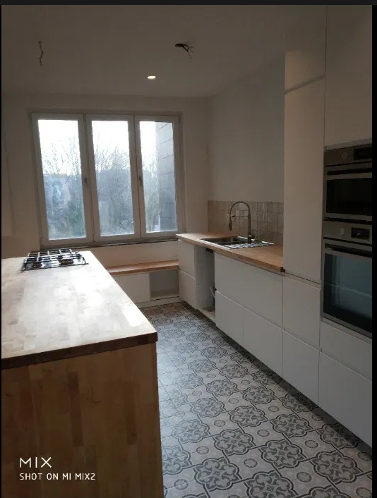 Jasna kuchnia z drewnianym blatem, białymi szafkami, wzorzystą podłogą i wnęką z siedziskiem pod oknem; widoczne niedokończone podłączenie oświetlenia sufitowego.