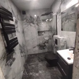 Wykończona łazienka z szarymi marmurkowymi płytkami na ścianach i podłodze, czarną armaturą i grzejnikiem drabinkowym, oświetleniem punktowym i zabudowaną półką prysznicową.